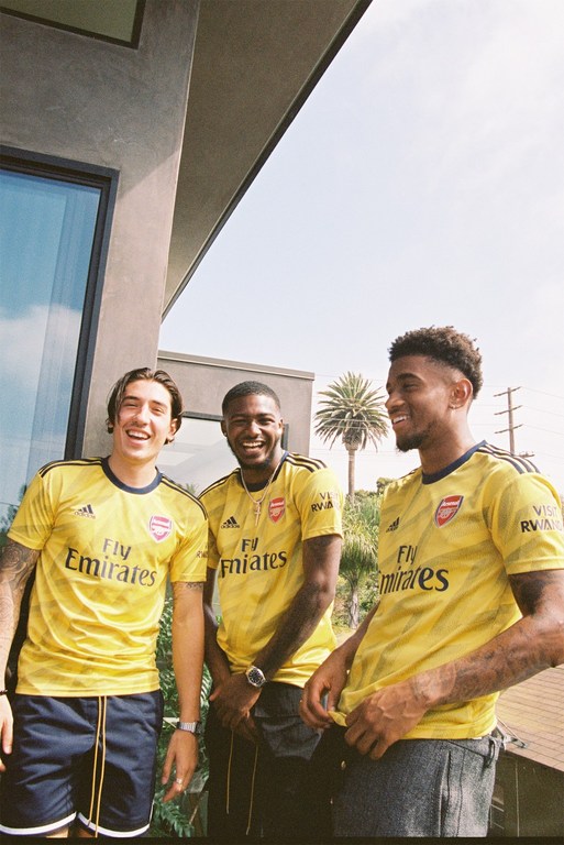 Three adult males smile outdoors wearing yellow Arsenal soccer jerseys, with a building and palm trees in the sunny background.