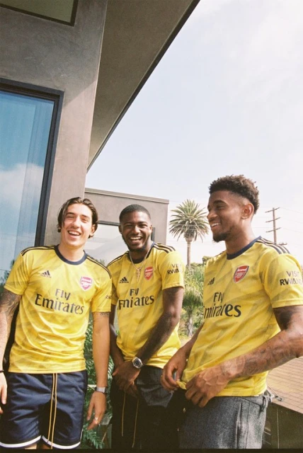 Three adult males smile outdoors wearing yellow Arsenal soccer jerseys, with a building and palm trees in the sunny background.