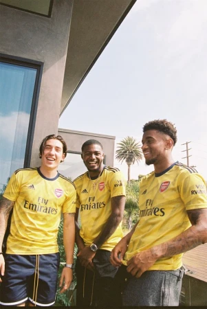 Three adult males smile outdoors wearing yellow Arsenal soccer jerseys, with a building and palm trees in the sunny background.