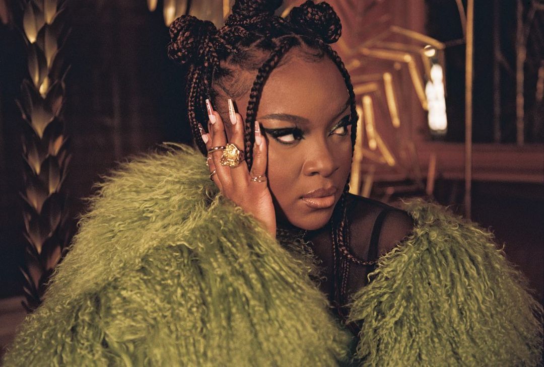 Close-up of a woman with an olive fur coat, elaborate braided hairstyle, and bold winged eyeliner posing indoors with a blurred background.