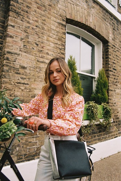A young woman with a bike stands in front of a brick building wearing an orange and white patterned top and white pants.