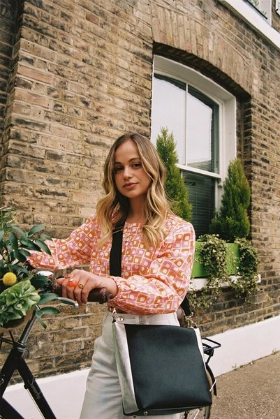 A young woman with a bike stands in front of a brick building wearing an orange and white patterned top and white pants.