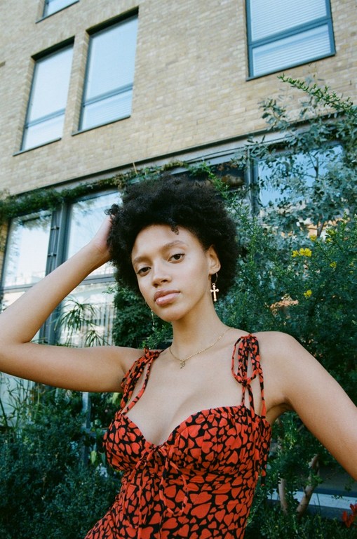 A young woman with dark curly hair poses in a red dress with black heart print in an outdoor urban setting with a brick building.