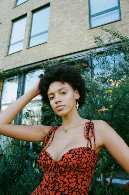 A young woman with dark curly hair poses in a red dress with black heart print in an outdoor urban setting with a brick building.