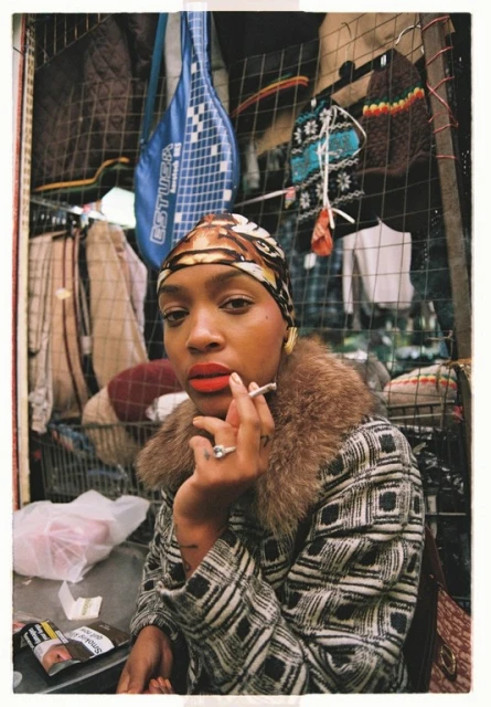A woman wearing a patterned headscarf and coat with a fur collar is smoking against a background of clothing and accessories.