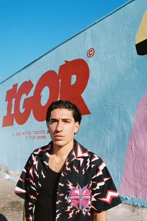 Adult male in black shirt and patterned button-up stands in front of a mural wall in light blue, red, pink, and yellow.