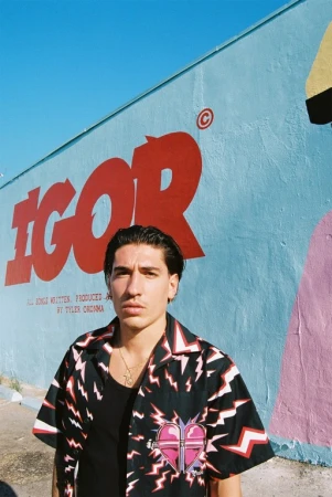 Adult male in black shirt and patterned button-up stands in front of a mural wall in light blue, red, pink, and yellow.