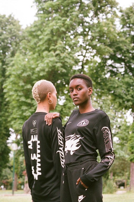 Two African-descent adult females pose outdoors in black long-sleeved shirts and black sweatpants with white graphics and lettering.