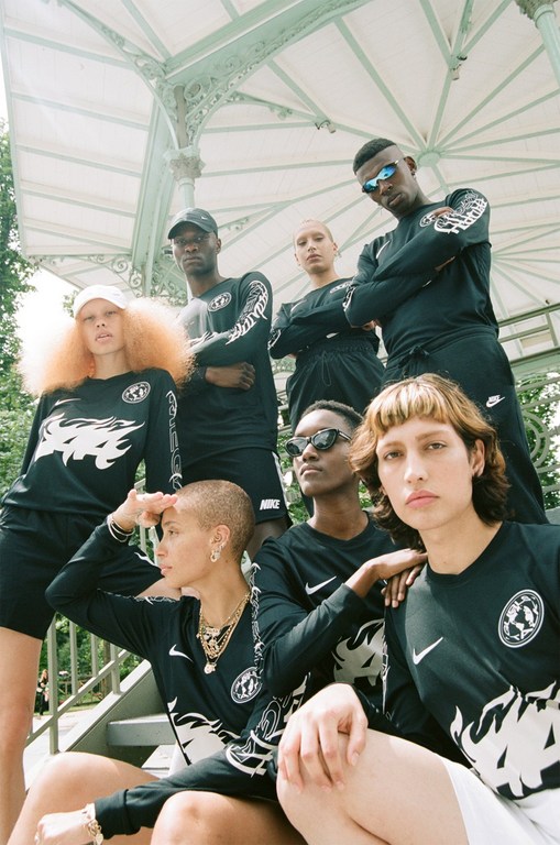 Group of seven diverse adults outdoors in black long-sleeve shirts with white flame graphics, posing on metal stairs with lush green trees.