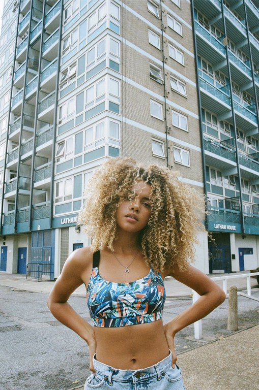 Woman with curly blond hair wears a patterned crop top and jeans against a tall city apartment building on a sunny day.
