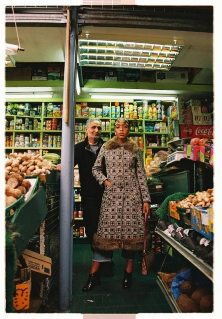 A fashionably dressed woman poses in a grocery store, with a man standing in the background amid aisles of products. 
