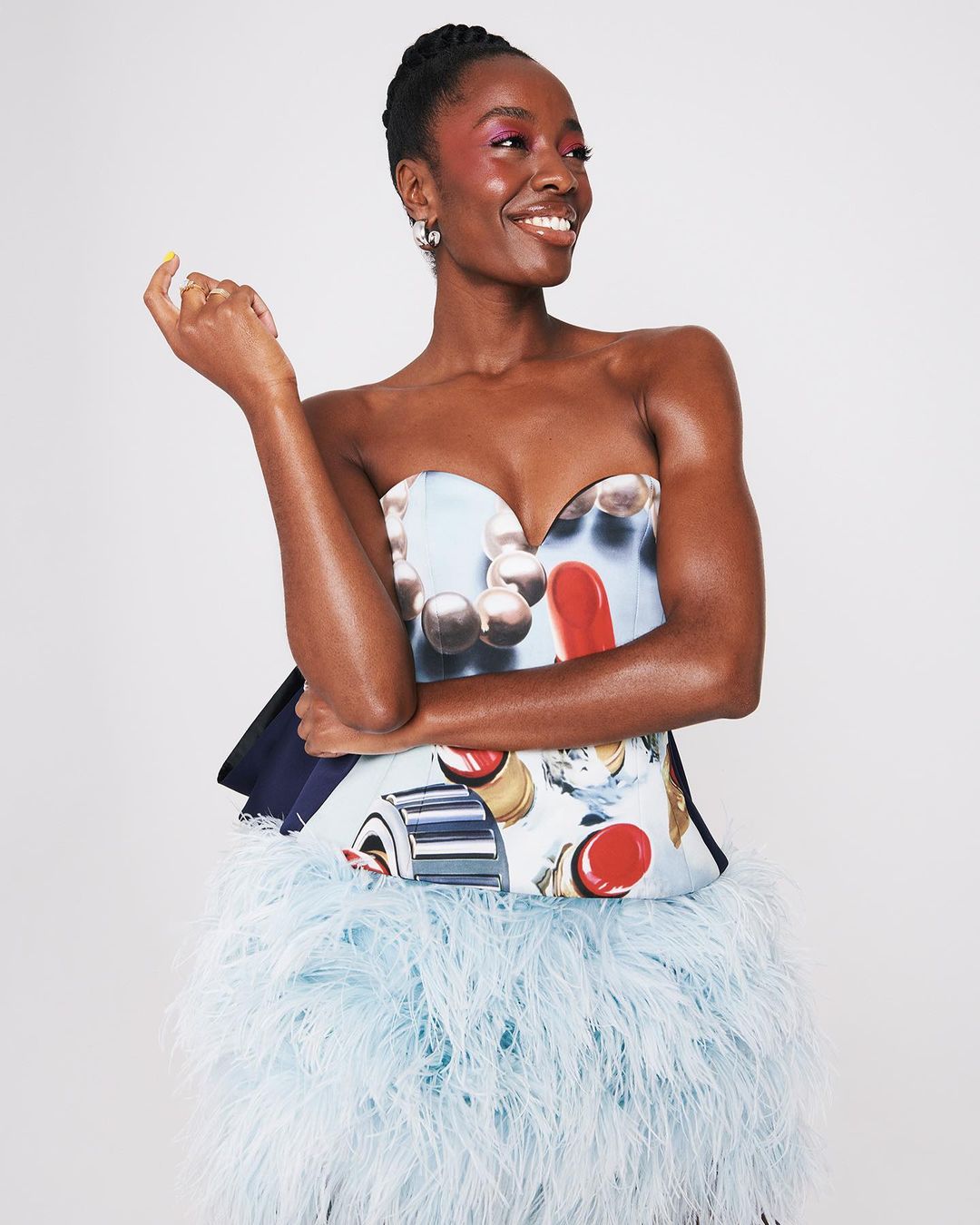 Smiling black woman in a strapless dress with pearl and lipstick print and a feather skirt in a studio portrait with a white background.