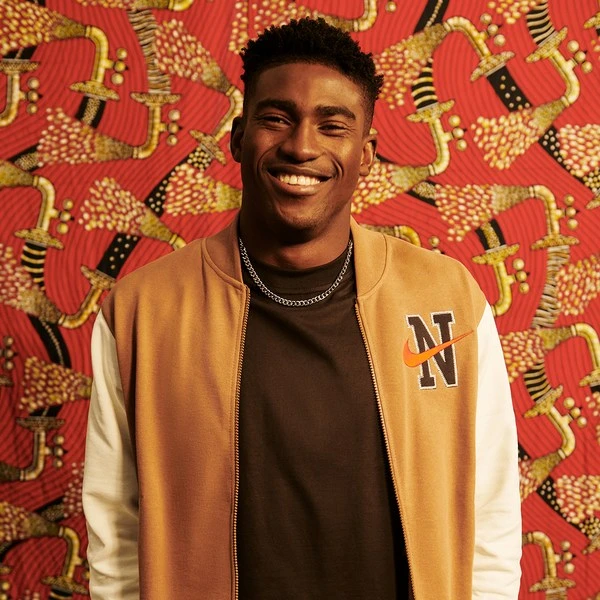 A smiling young Black male poses in a tan and white Nike bomber jacket in a studio with a vibrant patterned red backdrop.