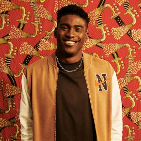 A smiling young Black male poses in a tan and white Nike bomber jacket in a studio with a vibrant patterned red backdrop.
