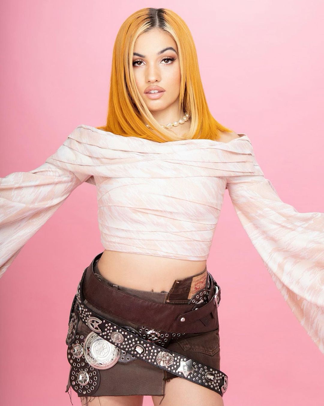 Portrait of a blonde woman in an off-the-shoulder top, embellished belt, and denim skirt against a light pink studio backdrop.  