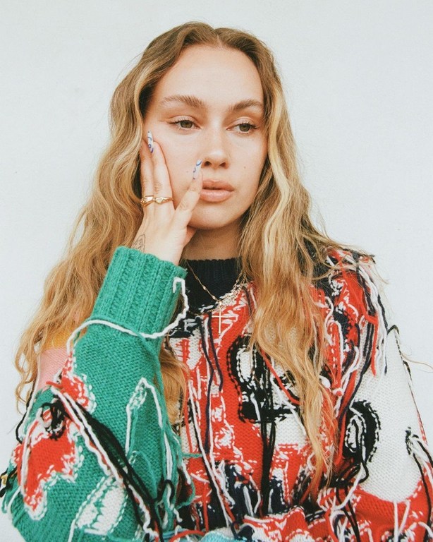 Close-up portrait of a woman with blonde highlighted hair wearing a colourful patterned sweater against a white background