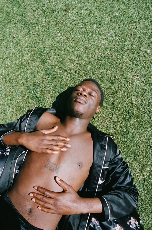 Man lies on grass in silk robe. An adult male with dark hair lays in sunlight with hands on his torso and a floral robe.