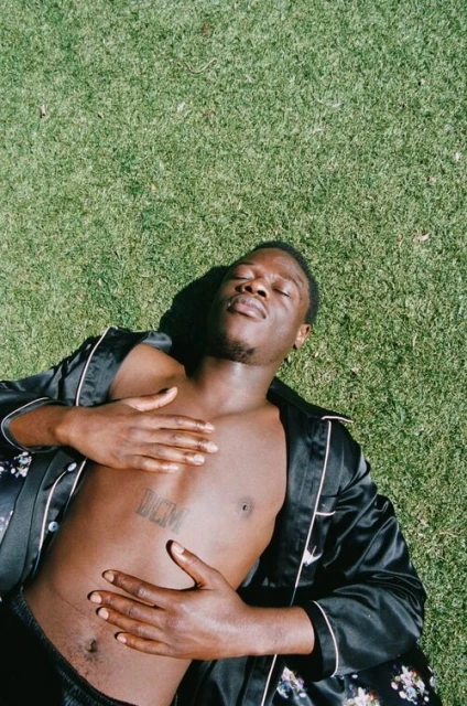 Man lies on grass in silk robe. An adult male with dark hair lays in sunlight with hands on his torso and a floral robe.