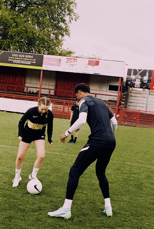 Female soccer player dribbles a soccer ball on a grassy field with a male player nearby and stadium seating in the background.