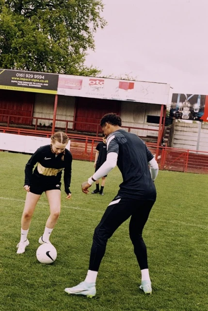 Female soccer player dribbles a soccer ball on a grassy field with a male player nearby and stadium seating in the background.