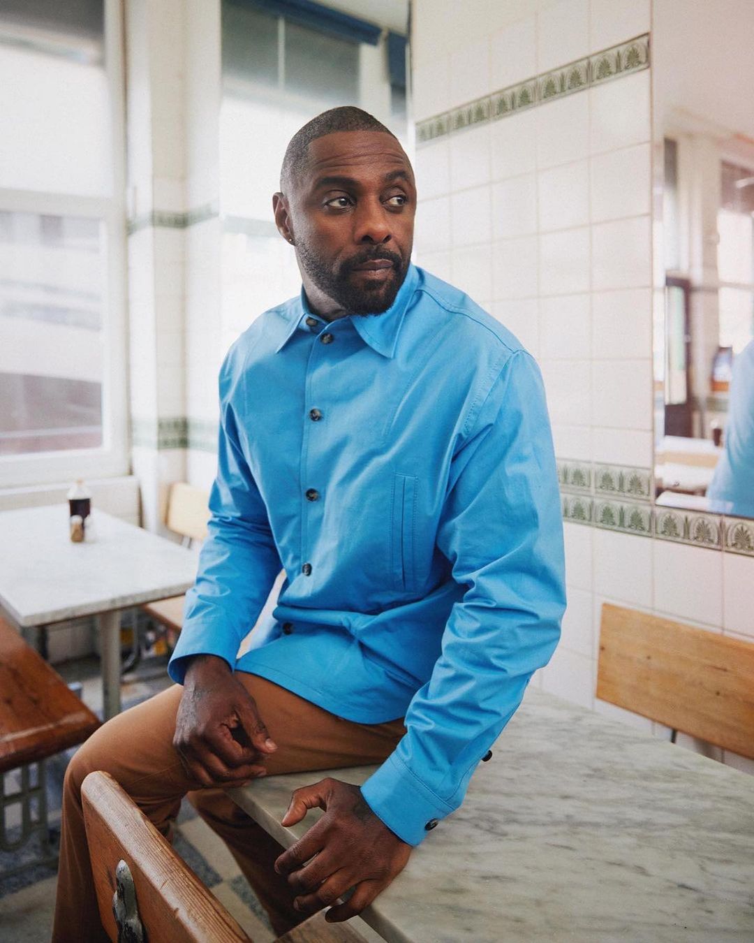Idris Elba poses indoors on a marble table wearing a blue button-down shirt and tan pants in a bright, tiled room with natural lighting.