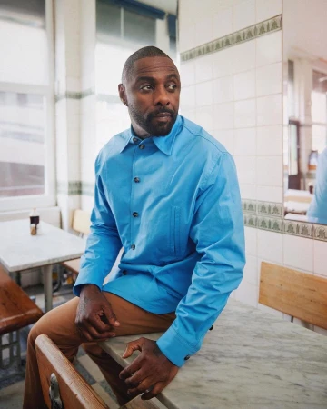 Idris Elba poses indoors on a marble table wearing a blue button-down shirt and tan pants in a bright, tiled room with natural lighting.