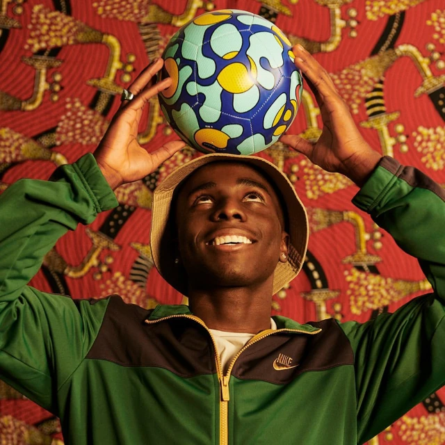 Young man balances a soccer ball on his head indoors. He wears a beige bucket hat, green jacket, and smiles against a patterned backdrop.