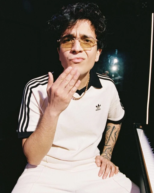 Portrait of a stylish adult with tattoos, wearing an Adidas track suit, gold chain and glasses, pouting, set against a dark background.