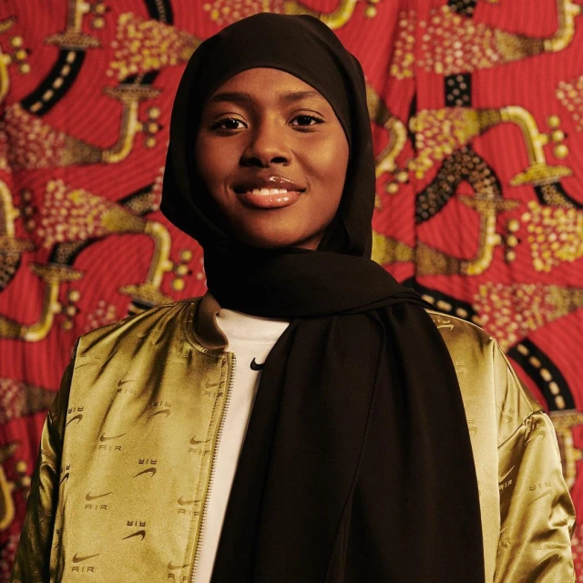 Close-up studio portrait of a smiling adult female wearing a black hijab, bomber jacket, and patterned backdrop with a medium shot.