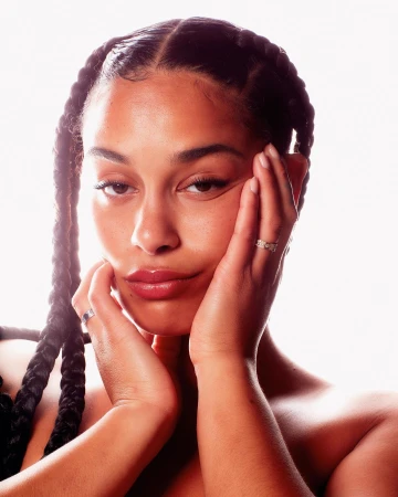 Close-up portrait of a woman with braided hair holding her face with both hands against a bright white background.