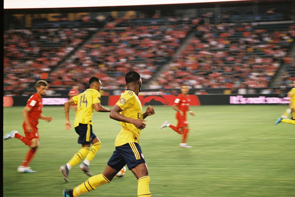 Soccer players in yellow and red uniforms compete on a green field during a match in a stadium filled with spectators.