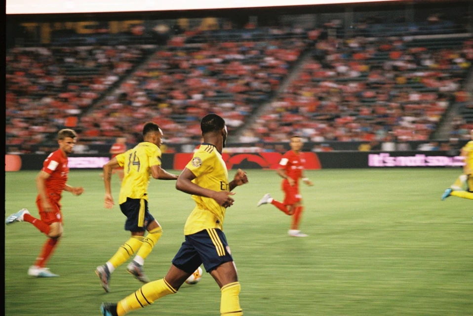 Soccer players in yellow and red uniforms compete on a green field during a match in a stadium filled with spectators.