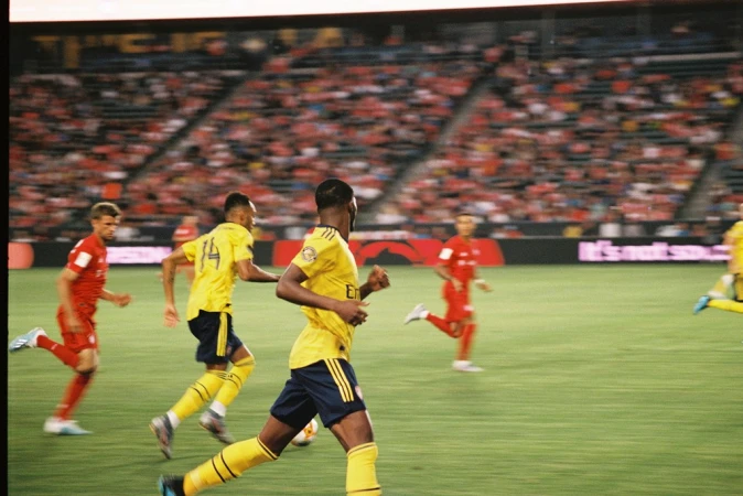 Soccer players in yellow and red uniforms compete on a green field during a match in a stadium filled with spectators.