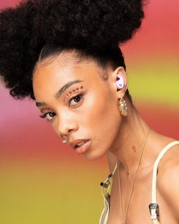 Close-up portrait of a woman with voluminous afro hair wearing purple wireless earbuds and gold hoop earrings against a gradient background.