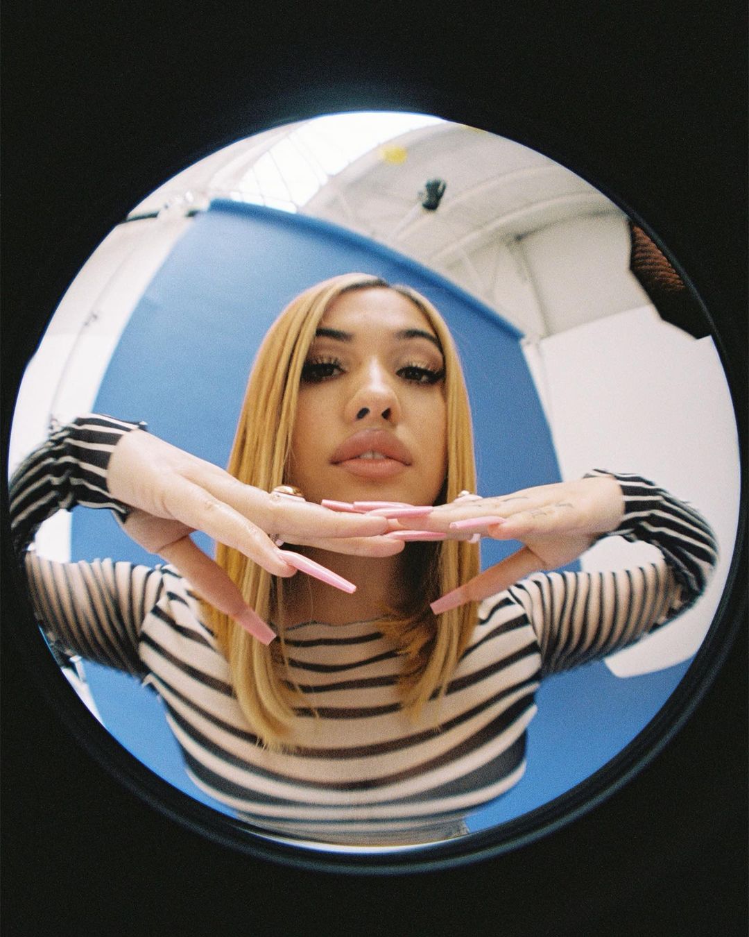 Portrait of a young adult female with blonde hair and long pink nails posing indoors in front of a blue background with studio lighting.