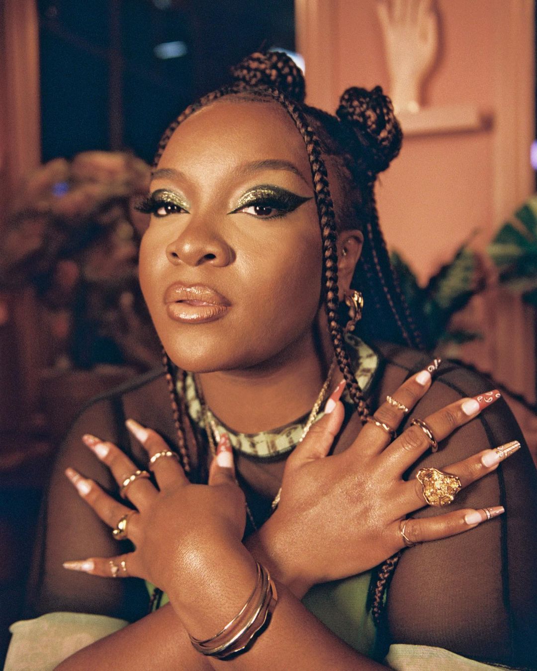 Portrait of a woman with braided hair, wearing gold jewelry, crossing her hands to show rings, green eye shadow, and long decorated nails.