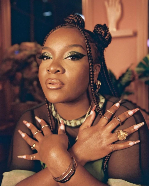 Portrait of a woman with braided hair, wearing gold jewelry, crossing her hands to show rings, green eye shadow, and long decorated nails.