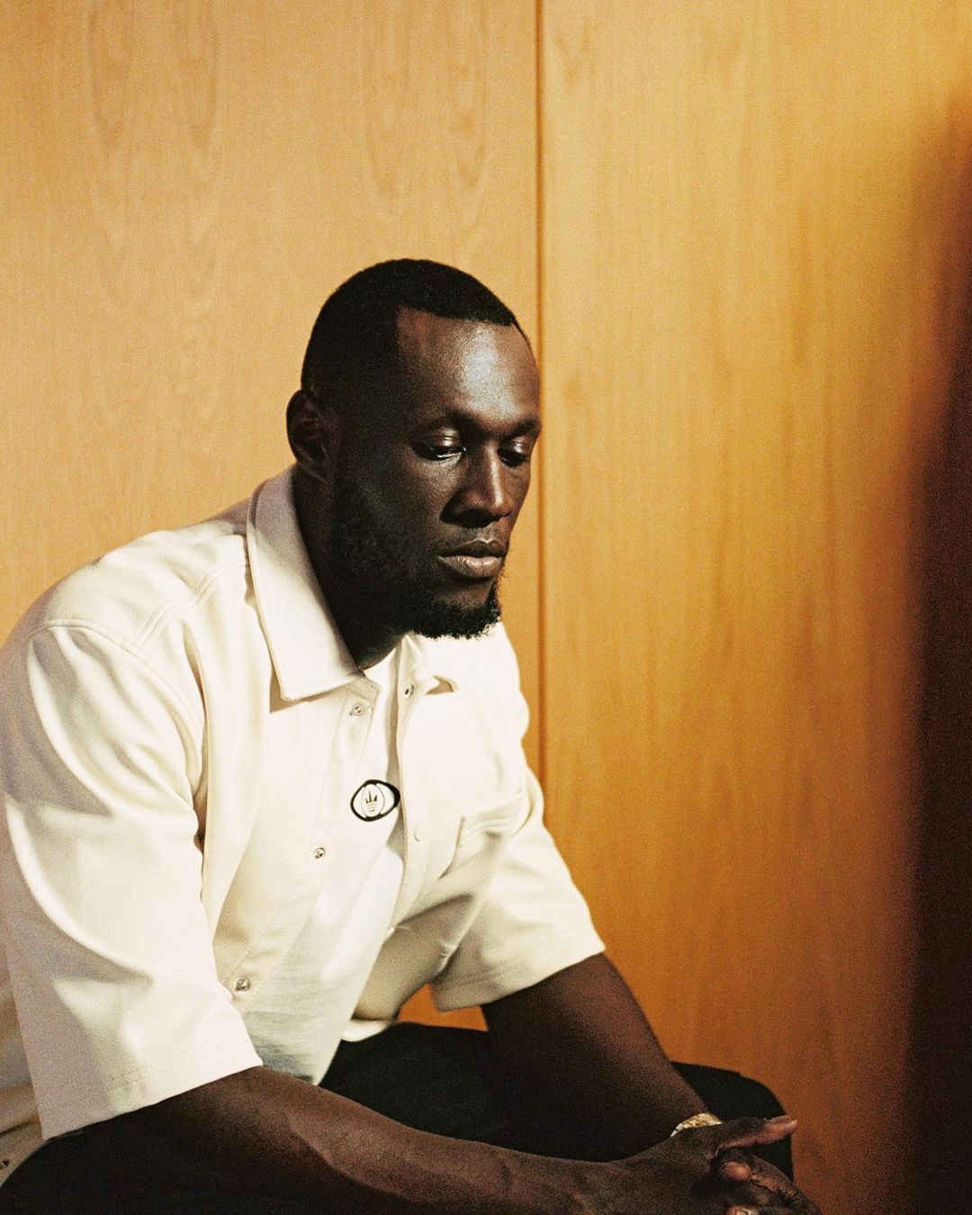 Studio portrait of African American man wearing a cream-colored work shirt and seated against a light-brown paneled wall.