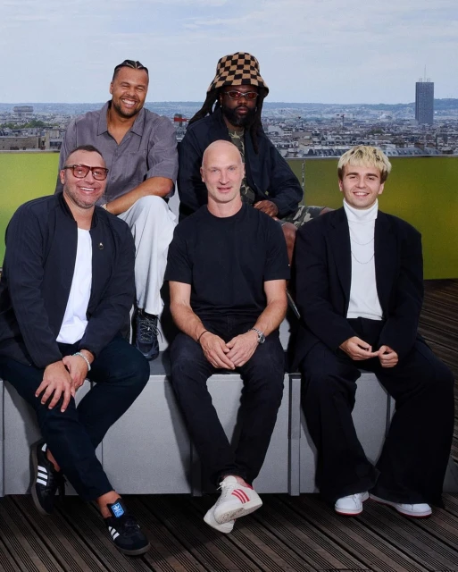 Group portrait of five men of diverse ethnicities seated outdoors with a cityscape background. They wear casual attire in a medium shot.