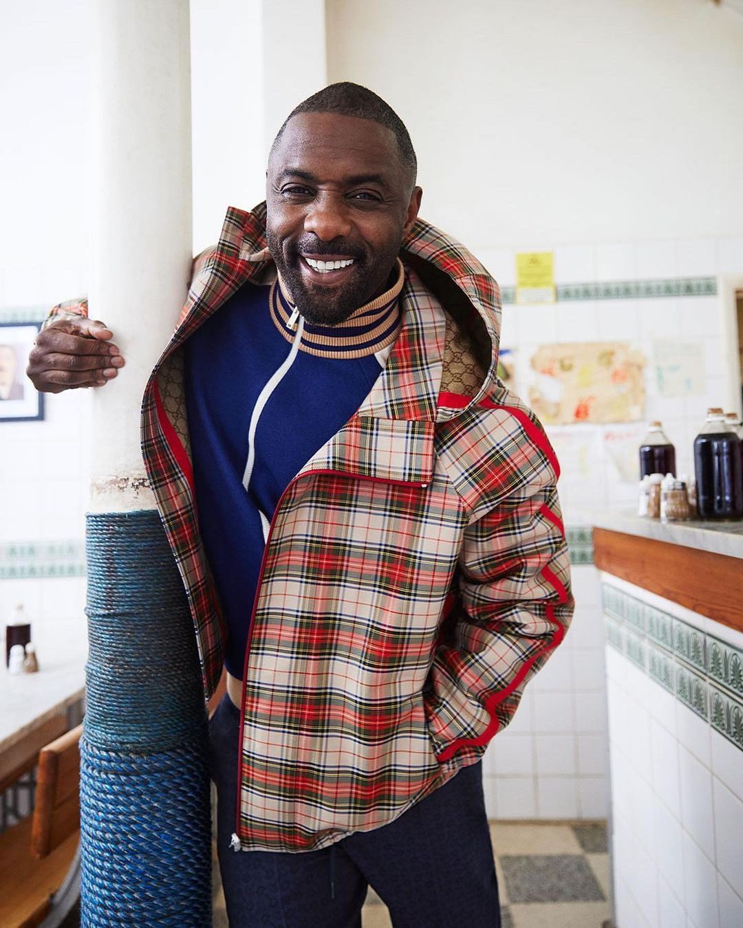 Idris Elba poses indoors wearing a plaid jacket and blue sweater. He leans against a white pillar with a smiling expression.