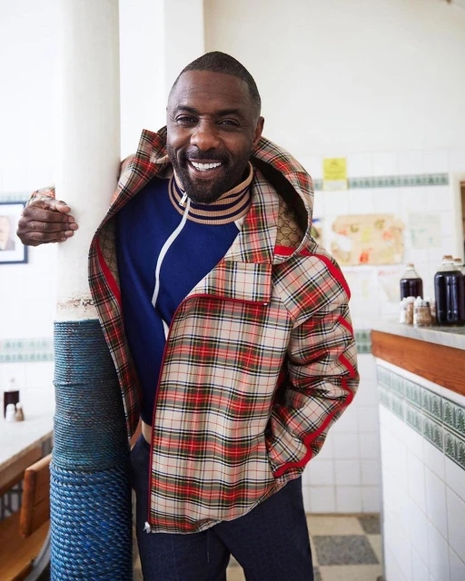 Idris Elba poses indoors wearing a plaid jacket and blue sweater. He leans against a white pillar with a smiling expression.
