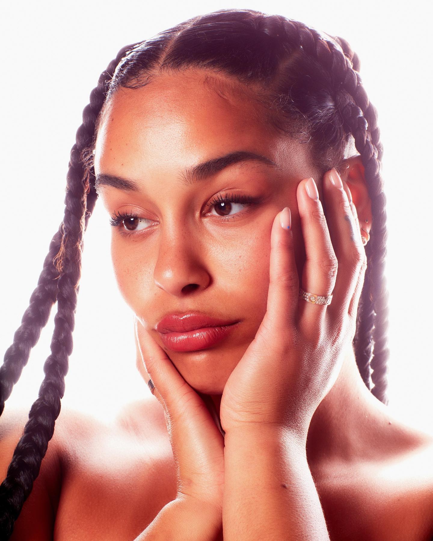 Close-up portrait of an adult woman with hands near her face, showcasing braided hair, glossy lips, and natural makeup against a bright background.