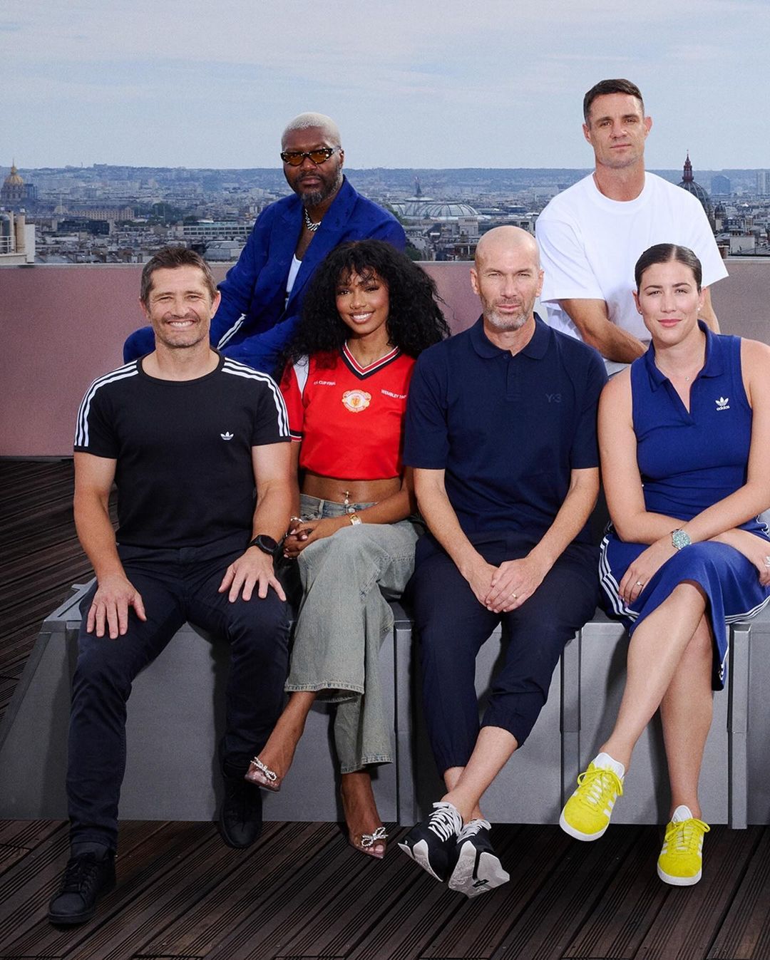 Group of six adults posing outdoors wearing Adidas apparel in an urban environment. Includes male and female adults and city view.