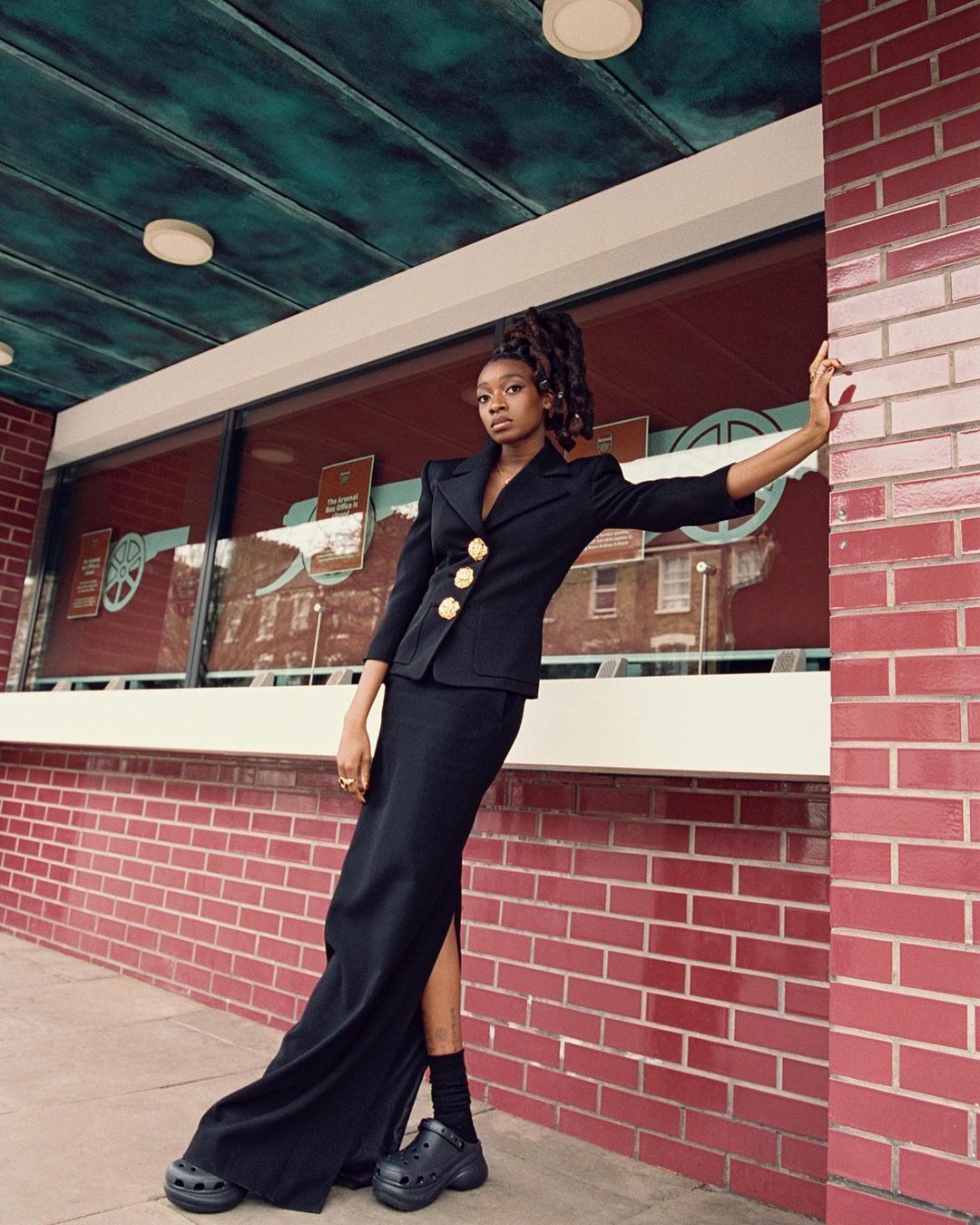 Elegant African American woman in a black blazer and skirt poses against a red brick wall, wearing black platform Crocs, fashionable and chic.