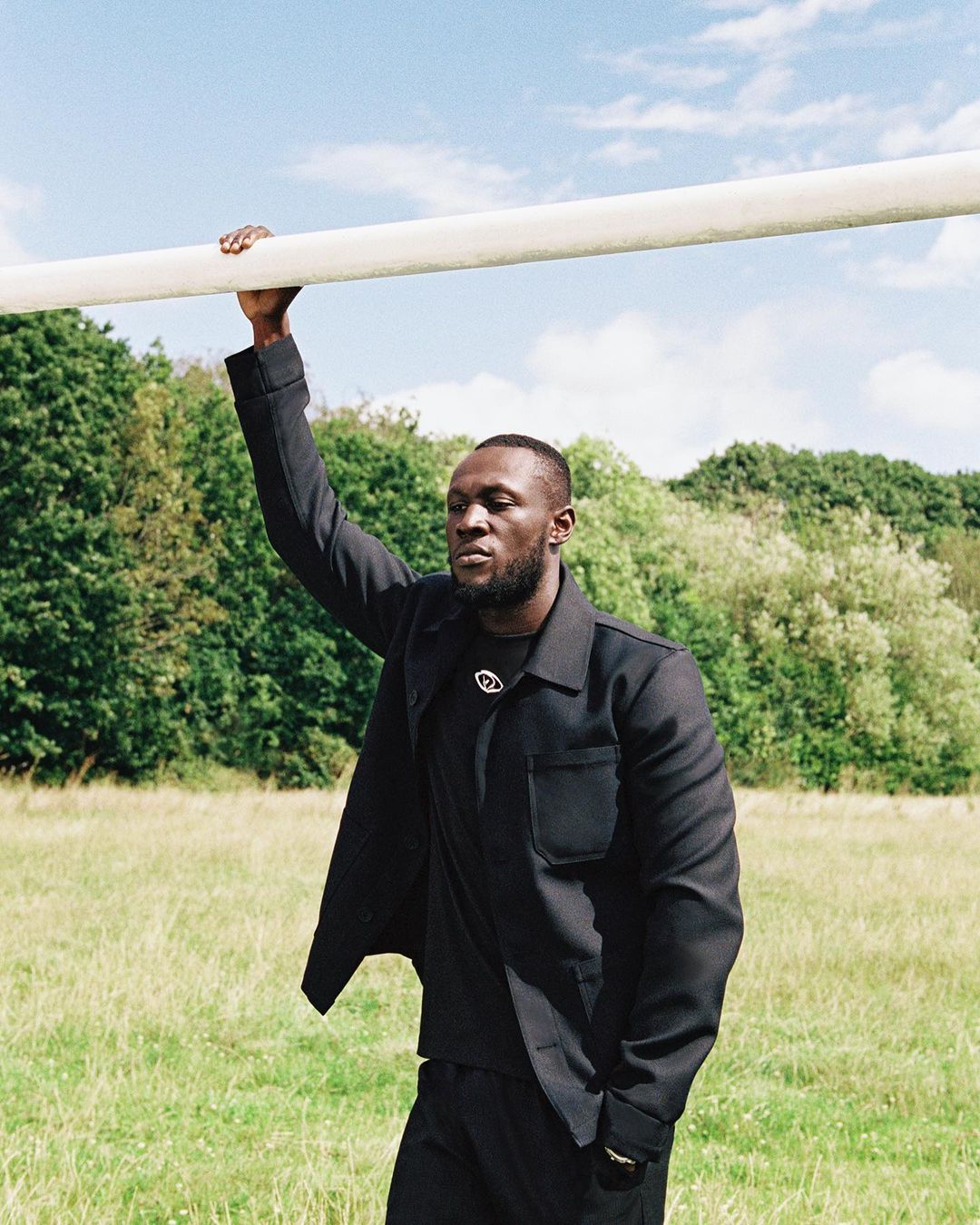 A Black adult male stands in a grassy field with trees, wearing a dark jacket and pants, holding onto a horizontal white bar overhead.