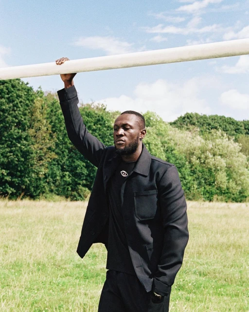 A Black adult male stands in a grassy field with trees, wearing a dark jacket and pants, holding onto a horizontal white bar overhead.