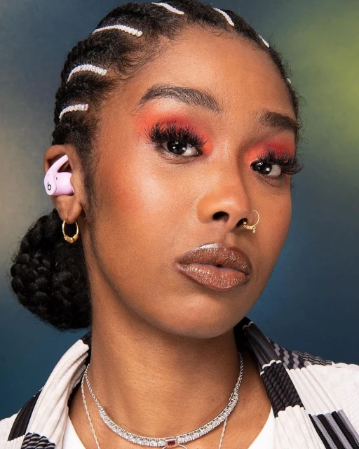 Close-up portrait of a stylish adult woman with pink earbuds, makeup, gold jewelry, and a patterned jacket against a blue and green backdrop.