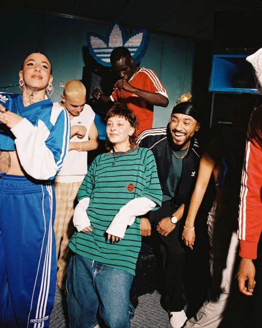 Group of young adults wearing Adidas clothing in an indoor setting with an Adidas logo on the wall, posing and smiling for a photo.