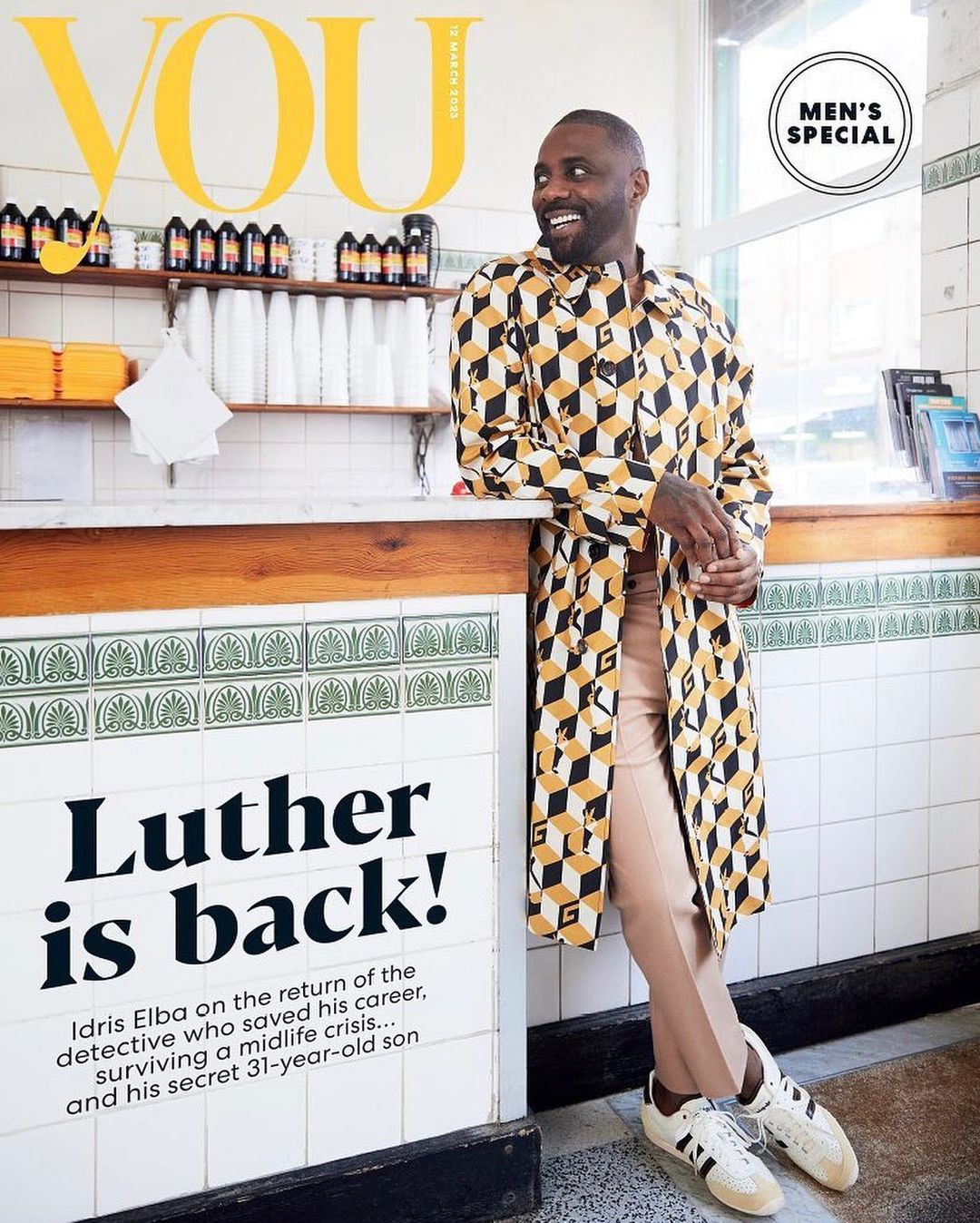 Magazine cover featuring Idris Elba in patterned trench coat and beige pants, standing against a white tile wall with vintage interior.