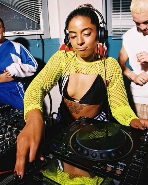 Young woman DJ in fishnet top plays music on a DJ deck, with two people in the background, in an indoor setting with window blinds.